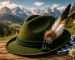Traditional green Lederhosen hat with feather and Edelweiss pin on a rustic wooden table against a scenic Bavarian Alpine background.